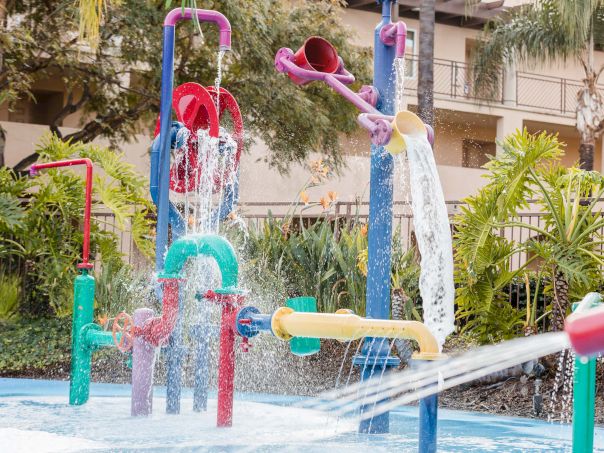 A colorful splash pad with water sprayers and fountains shooting water in multiple directions, surrounded by plants and a building in the background.