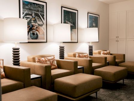 A cozy waiting room with tan armchairs, matching ottomans, floor lamps, framed travel photos, and a white cabinet in the corner.